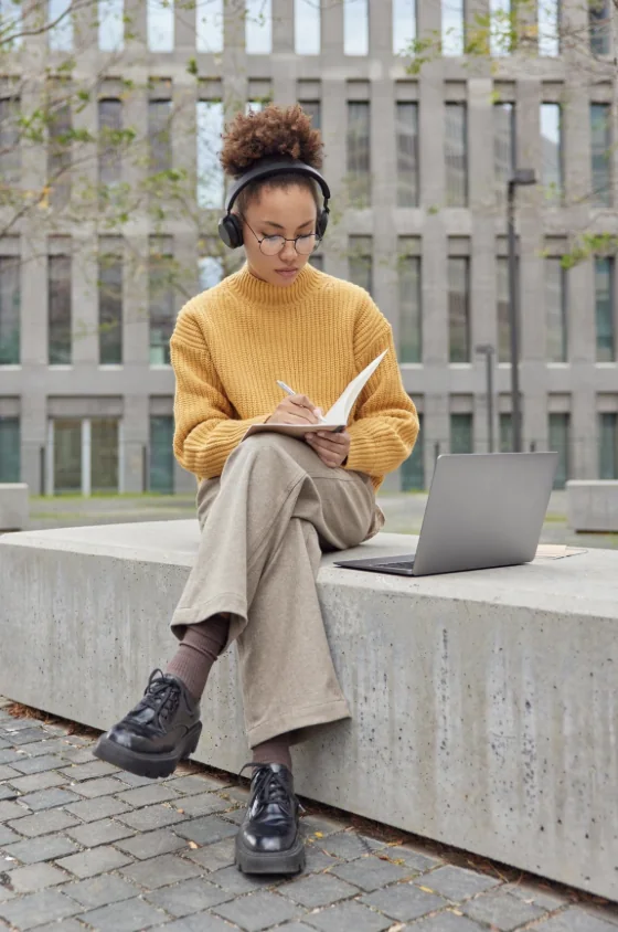Une apprenante prend des notes dans un carnet, casque sur les oreilles et ordinateur à côté.